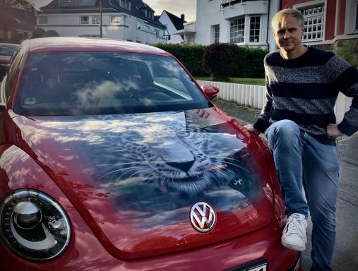 Mann steht lässig neben einem roten Volkswagen Käfer mit Wolkenmuster auf der Motorhaube.